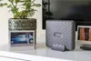 A Fujifilm Instax LINK WIDE printer is displayed on a white surface next to a potted plant and a framed photo. Produktbild-Vorschau