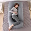 A pregnant woman is lying on a bed, using a J-shaped pregnancy pillow for support. Náhled obrázku produktu