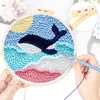 A person's hands are shown working on a punch needle embroidery kit featuring a whale and ocean scene. Produktbild-Vorschau