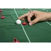 A hand is playing a tabletop soccer game, moving a white ball with a finger. Miniature soccer players are positioned on a green field with white lines. Produktbild-Vorschau