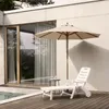 A white plastic sun lounger and a beige umbrella are set up on a wooden deck next to a pool, with a modern building in the background. Voir les images du produit