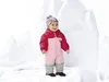 A baby wearing a pink and red snowsuit and a patterned hat stands in a snowy environment with large ice formations in the background. Produktbild-Vorschau