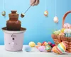 A pink chocolate fountain is shown, with a hand dipping a marshmallow into it. There are hanging Easter eggs and a basket of Easter eggs with flowers in the foreground. Náhľad obrázku produktu