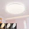 A modern LED ceiling light with a star-like pattern is shown mounted on a white ceiling in a room with pink walls. Below the main light, three smaller circular swatches display different light color temperatures. A white remote control is visible in the lo Produktbild-Vorschau