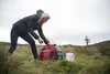 A woman is operating a red Honda generator in a grassy, outdoor environment. Produktbild-Vorschau