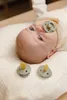 A baby lying on a cream-colored blanket with two pacifiers in front of them and one in their mouth. Produktbild-Vorschau
