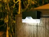 A close-up of a black LED solar light attached to a wooden fence, with blurred greenery in the background. Podgląd zdjęcia produktu