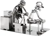 A metal sculpture of two figures, one appearing to be a tattoo artist working on another figure, with a toolbox and equipment nearby. Produktbild-Vorschau