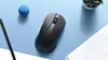 A black wireless mouse is placed on a light blue surface, next to a pencil, a blue ball, and a small potted plant. A white document is partially visible in the foreground. Voir les images du produit