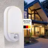 A modern outdoor LED light fixture with a motion sensor is mounted on a textured wall, with a blurred background showing a contemporary house at dusk. Produktbild-Vorschau