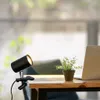 A black clip-on desk lamp is attached to a wooden table next to a potted plant and a laptop. Náhled obrázku produktu