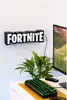 A Fortnite logo light is mounted on a white wall above a desk with a computer monitor, keyboard, and plant. Anteprima dell’immagine prodotto
