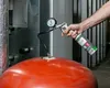 A hand holding a can of spray and a pressure gauge connected to a red expansion tank. Produktbild-Vorschau