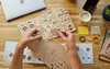Hands assembling a wooden 3D puzzle on a wooden desk with a laptop, coffee, and other small items. Anteprima dell’immagine prodotto