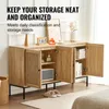 A wooden sideboard with rattan accents, featuring open doors revealing shelves and storage, set in a room with a lamp, calendar, and decorative items on top. Anteprima dell’immagine prodotto
