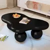 A black cloud-shaped coffee table with spherical legs is placed on a plush rug. There's a bowl of fruit and a cup of coffee on the table, and an open magazine on the floor. Voir les images du produit