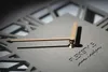 A close-up shot of a modern wall clock with Roman numerals and a minimalist design. The clock hands are made of wood and gold-toned metal. Náhled obrázku produktu