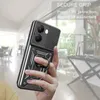 A hand holding a black smartphone case with a ring holder and camera cover, in a blurred indoor setting. Produktbild-Vorschau