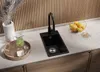 A black kitchen sink with a black faucet is shown on a countertop with a bowl of fruit and a vase of dried flowers. Produktbild-Vorschau