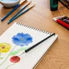 A close-up of a sketchbook with watercolor paintings of flowers, surrounded by art supplies like paintbrushes and tubes of paint on a wooden table. Produktbild-Vorschau