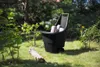 A black garden cart filled with logs is placed on a grassy lawn in a garden setting. Náhled obrázku produktu