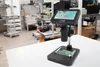 A digital microscope is set up on a white table in a laboratory or workshop setting, with a circuit board placed on its stage. Produktbild-Vorschau