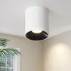 A white cylinder-shaped ceiling light with a black interior is mounted on a white ceiling. A green plant is visible in the lower left corner. Produktbild-Vorschau