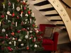 A decorated Christmas tree with lights and ornaments stands next to a red armchair and a staircase. Produktbild-Vorschau