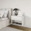A white floating nightstand with a drawer and an open shelf, placed next to a bed. Produktbild-Vorschau