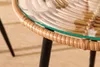 Close-up of a round glass-top table with a rattan edge and black legs, on a light-colored carpet. Produktbild-Vorschau