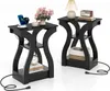 Two black side tables are shown against a white background. The tables have a modern, curved design and feature multiple shelves. The table on the left has a phone and smartwatch charging on its top surface, along with a cup and a vase of flowers. The tabl Produktbild-Vorschau
