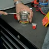 A person's hands are shown opening a can of NIGRIN Performance Fein-Spachtel with a screwdriver on a dark gray workbench. Other car detailing tools and supplies are visible in the background. Anteprima dell’immagine prodotto