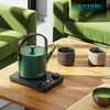 A green electric kettle with a brown handle sits on a black base on a wooden table, next to two textured cups. In the background, a green sofa is visible. Podgląd zdjęcia produktu