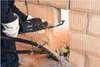 A person is using a reciprocating saw to cut through a brick wall. There are pipes and debris on the ground. Voir les images du produit