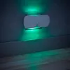 A white LED night light with motion sensor is mounted on a gray textured wall above a gray baseboard and wooden floor. Green light emanates from the night light, illuminating the wall and floor. Produktbild-Vorschau
