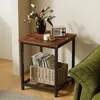 A square side table with a wood-look top and black metal frame, decorated with a vase of flowers, a candle, a small radio, and a basket of records on the shelf below. A green sofa is partially visible on the right. Produktbild-Vorschau