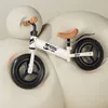 A white balance bike with a brown seat and handlebars is positioned on a beige, abstract background. Náhled obrázku produktu