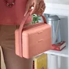 A person is holding a pink lunchbox with a strap. The lunchbox is on a shelf with books and a watering can. Podgląd zdjęcia produktu