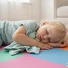 A young child is sleeping on a colorful puzzle mat, holding a stuffed toy. Produktbild-Vorschau