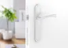 A white door handle and lock mechanism are shown in the foreground, with a blurred interior scene in the background. Produktbild-Vorschau