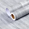 Two rolls of self-adhesive wood-effect wallpaper in a gray wood grain pattern are shown diagonally across a surface with the same pattern. Náhľad obrázku produktu