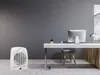 A white fan heater is placed on a concrete floor in front of a gray wall, next to a modern office desk with a computer and a chair. Produktbild-Vorschau