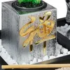 A close-up of a zen garden decoration with a silver cube featuring a gold symbol, surrounded by white pebbles and a bamboo stick. Náhled obrázku produktu