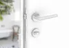 A close-up view of a modern door handle and lock mechanism on a white door, with a blurred bathroom scene visible through the open doorway. Produktbild-Vorschau