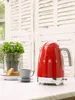 A red Smeg kettle is on a countertop next to a cup of coffee and some biscuits, with a plant in the background. Produktbild-Vorschau