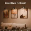 Drei schwarze verstellbare Bildleuchten, die gerahmte Kunstwerke über einem beigen Sofa mit Kissen beleuchten Náhled obrázku produktu