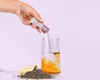 A hand is pouring powder from a sachet into a glass of water, next to a pile of loose tea leaves. Produktbild-Vorschau