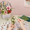 A person's hands are assembling a 3D wooden flower model kit, with a completed flower basket in the background. Podgląd zdjęcia produktu