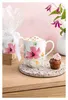 A floral mug filled with a frothy drink, a cupcake, and a vase of flowers in the background. Podgląd zdjęcia produktu
