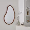 An organic-shaped mirror with a wooden frame hangs on a light gray wall next to a white shelf with decorative items. Anteprima dell’immagine prodotto